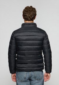Sort pufferjakke med vandret quiltet design, høj krave og ribbede manchetter, båret over lyseblå jeans. Glat tekstur, letvægts.
