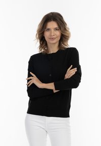 Cardigan negru, cu mâneci lungi, guler rotund, cu închidere cu nasture și manșete cu nervuri. Puratat peste pantaloni albi. Textură netedă.