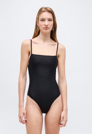 Kobieta o jasnej karnacji i długich blond włosach ubrana w prosty, czarny jednoczęściowy strój kąpielowy z cienkimi ramiączkami, stoi na tle szarego tła.