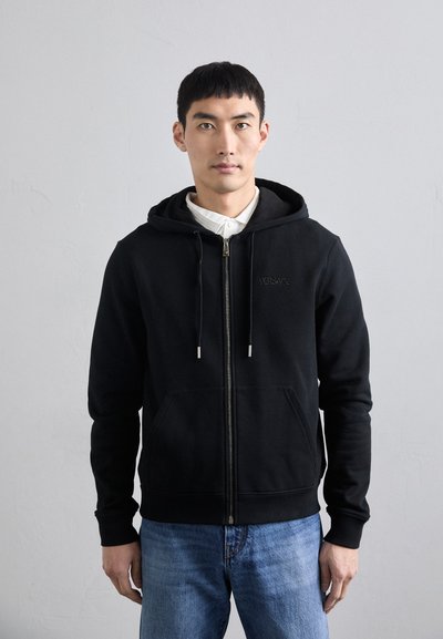 Schwarze Kapuzenjacke mit Reißverschluss, Kordelzugkapuze, Fronttaschen und subtilen Logodetails. Getragen über einem weißen Hemd mit Kragen, kombiniert mit blauen Jeans.
