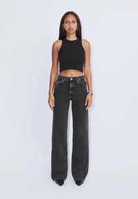 Kobieta stojąca przodem, ubrana w czarny, bezrękawnikowy crop top oraz czarne, szerokie spodnie dżinsowe z wysokim stanem i czarne, spiczaste buty.