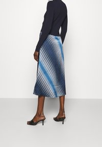 Kvinna iförd en marinblå långärmad topp och en midi kjol med zigzag-mönster i blått, vitt och svart, samt svarta klacksandaler.