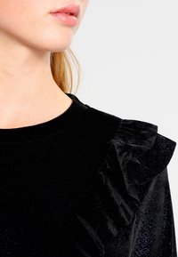 Svart sammetstopp med rund halsringning och volangdetaljer vid axlarna, med en mjuk textur och diskret glans.