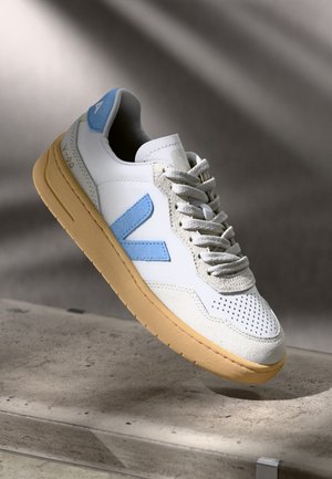 Biały sneaker z niebieskim logo "V", beżowymi detalami z zamszu, brązową podeszwą i białymi sznurówkami unoszący się nad betonową powierzchnią z delikatnymi cieniami w tle.