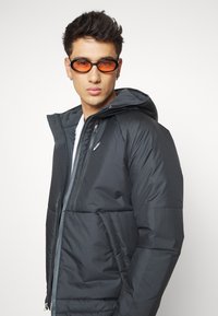 Dark padded jacket with a hood, featuring a zippered front and side pockets. Gray interior lining. Model wears orange-tinted sunglasses.