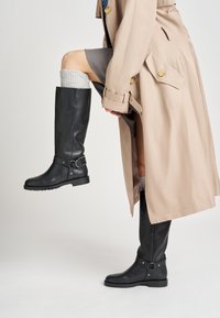 Botas negras de cuero hasta la rodilla con un acento de hebilla, acabado texturizado y un tacón bajo. Llevadas con un abrigo trinchera de color beige claro.