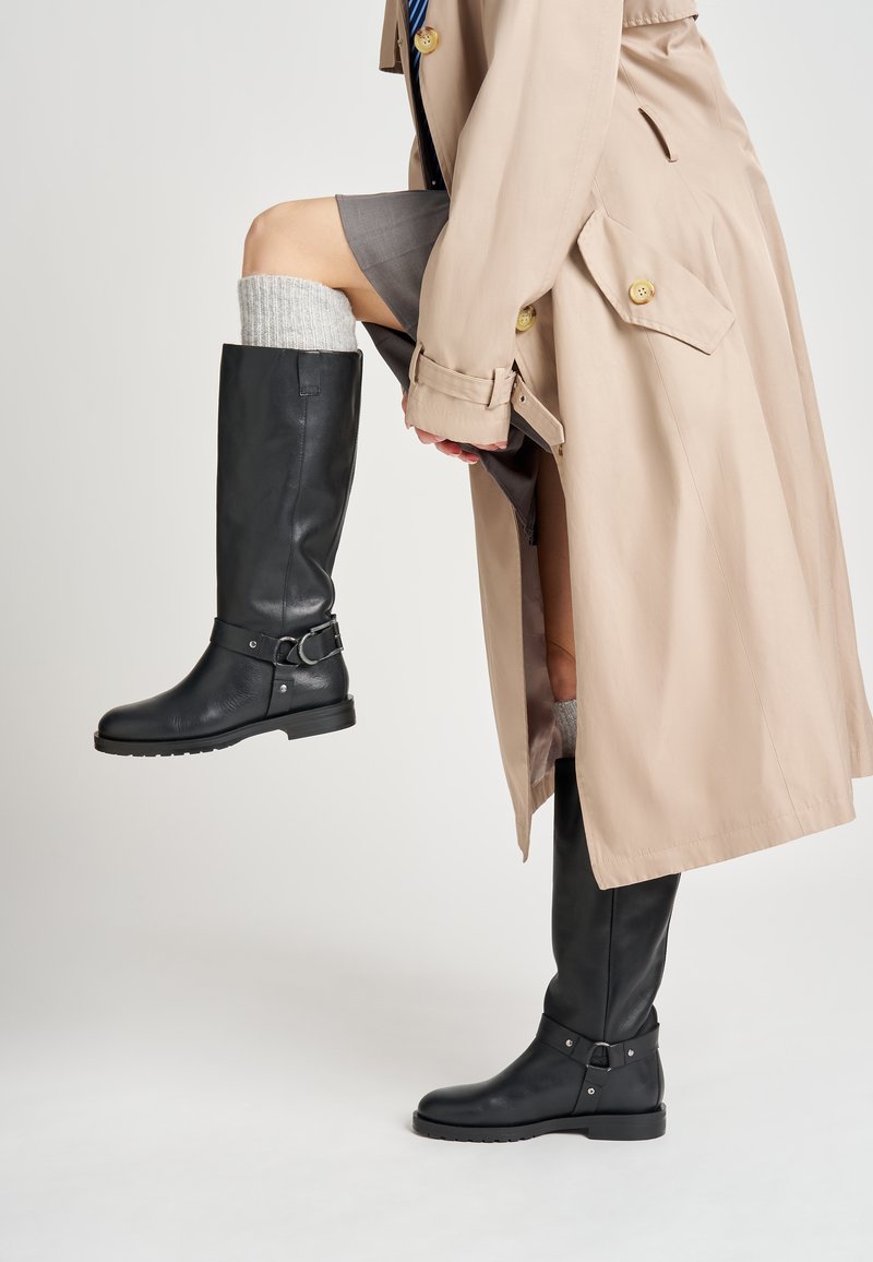 Botas negras de cuero hasta la rodilla con un acento de hebilla, acabado texturizado y un tacón bajo. Llevadas con un abrigo trinchera de color beige claro.