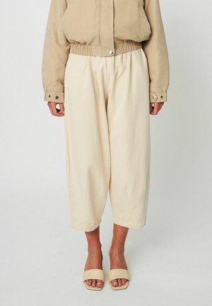 Beige Caprihose mit elastischem Bund, getragen zu einer passenden beigen Cropped-Jacke und beigen, offenen Sandalen mit Riemchen.