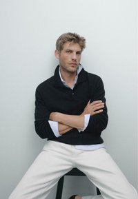 Schwarzer Strickpullover, der über einem hellblauen gestreiften Hemd getragen wird, kombiniert mit weißen Hosen. Die Person sitzt auf einem schwarzen Stuhl, die Arme verschränkt.