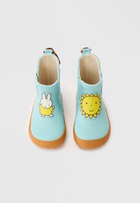 Bottes de pluie en caoutchouc bleu avec des semelles brunes. La botte gauche présente un graphique de soleil jaune ; la botte droite a un dessin de lapin en robe jaune.