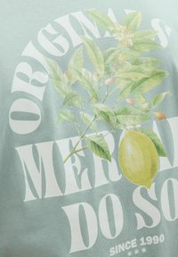 Mintgrøn T-shirt med et grafisk motiv af citronbrancher med grønne blade og lyserøde knopper, kombineret med fed hvid tekst.
