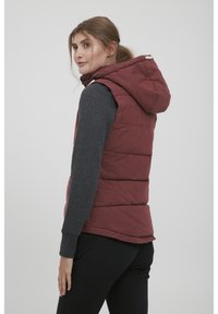 Gilet matelassé rouge avec capuche, doté d'une fermeture éclair et de boutons-pression. Le gilet est superposé à une chemise à manches longues gris foncé.