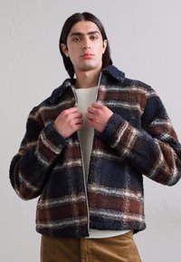 Casaco de fleece em padrão xadrez castanho e azul marinho, com gola e fecho de correr, combinado com um suéter texturizado em creme por baixo.