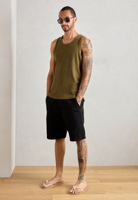 Alyvuogių spalvos juostelėmis marškinėliai, juodi šortai ir šviesūs sandalai. Asmuo turi tatuiruotes ant vienos rankos ir nešioja akinius nuo saulės.