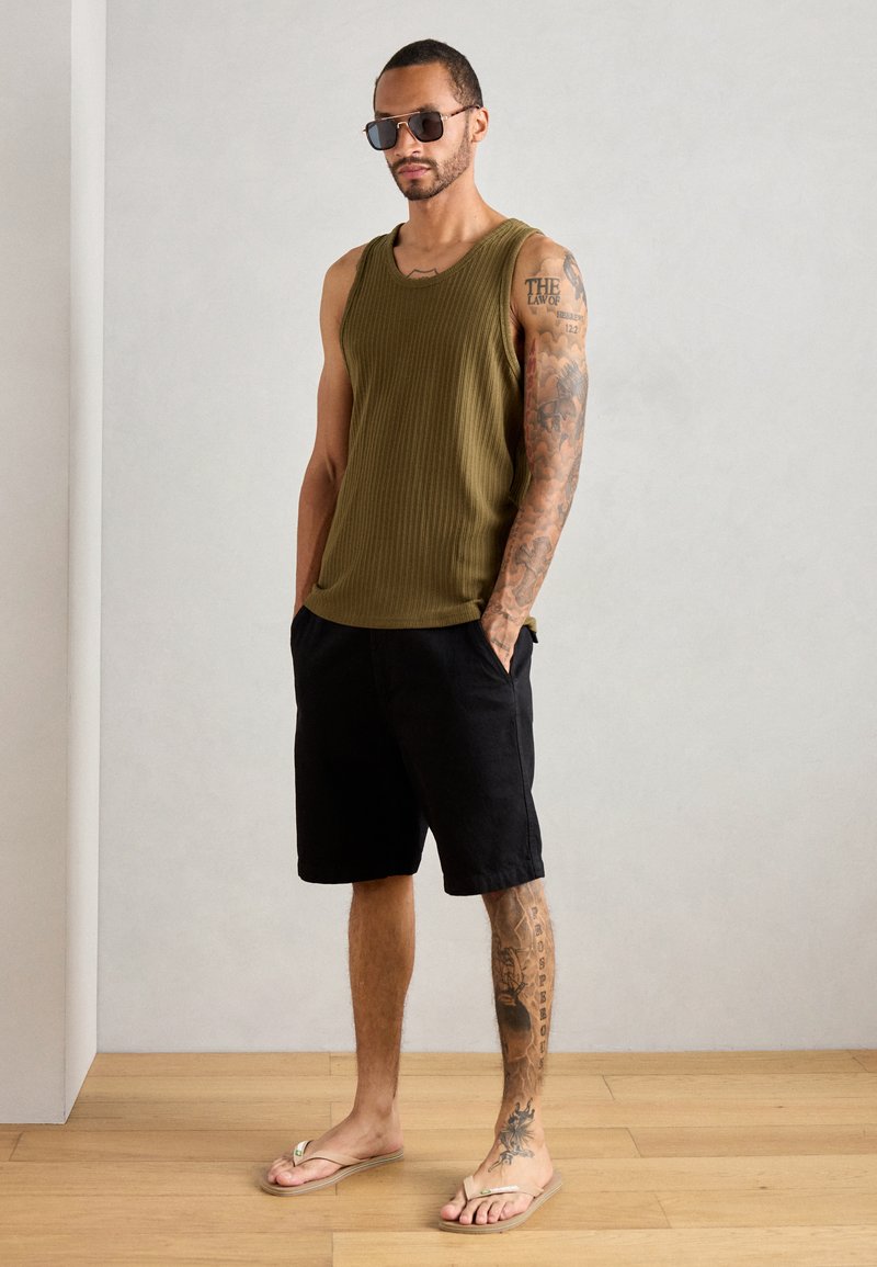 Débardeur à rayures olive, shorts noirs et sandales légères. L'individu a des tatouages sur un bras et porte des lunettes de soleil.