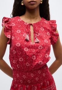 Femme portant une robe rouge à fleurs avec des manches à volants et un décolleté avec une ouverture en forme de goutte, attachée par des cordons à pompons.