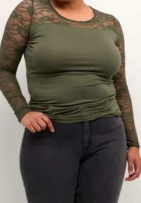 Oliivinvihreä, pitkähihainen paita, jossa pitsikoristeita harteilla ja hihoissa, istuva malli ja pehmeä koostumus, yhdistetty tummiin farkkuihin.