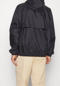 Svart, lättviktsjacka med dragsnodd i huvan, elastiska ärmslut och fåll. Har en bakflik för ventilation. Bärs över beige byxor.