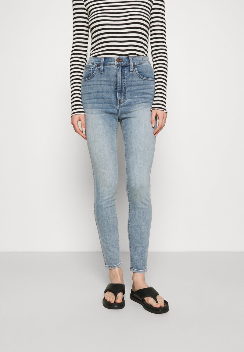 Personne portant un jean skinny taille haute bleu clair, des sandales noires à enfilage et un haut à rayures noir et blanc à manches longues, adossée à un mur blanc.