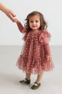 Rynket lyserød kjole med gyldne bue-mønstre over en ensfarvet langærmet top. Inkluderer beige strømpebukser og mørkegrønne sko med spænder.