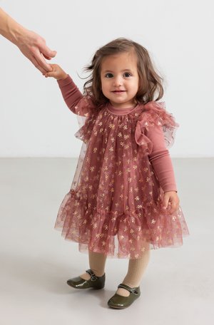 Robe rose froncée avec des motifs de nœuds dorés sur un haut à manches longues uni. Comprend des collants beiges et des chaussures vert foncé avec des boucles.