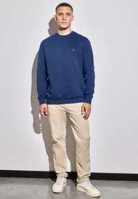 Mörkblå sweatshirt med långa ärmar och rund halsringning, i kombination med beiga byxor och vita sneakers, mot en neutral bakgrund.