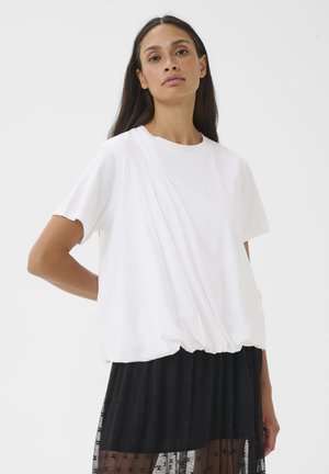 Femeie cu păr lung și închis la culoare, purtând un top alb lejer cu mâneci scurte și tiv adunat, și o fustă neagră transparentă cu modele florale.