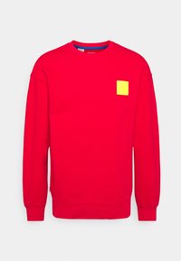 Rode longsleeve sweatshirt met ronde hals en ribbels aan de manchetten, met een klein geel vierkant embleem dat textuur heeft zoals Lego-blokken op de linkerborst.