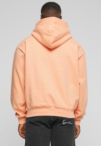 Sudadera en melocotón claro con capucha ajustable, puños y dobladillo acanalados. Presenta un acento de logo en el bolsillo trasero de unos jeans negros.