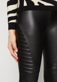 Svarta läderleggings med en slät yta, med horisontell ribbad detalj längs sidan. Materialet ser snyggt och åtsittande ut.