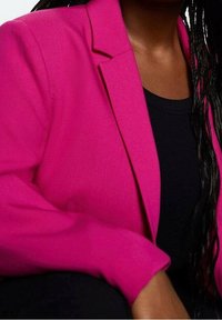 Personne portant un blazer rose vif sur un haut noir, avec de longues tresses visibles sur un côté.