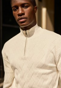 Suéter cremoso de cremallera con un patrón de diamante texturizado y cuello acanalado. La tela parece suave y de punto.