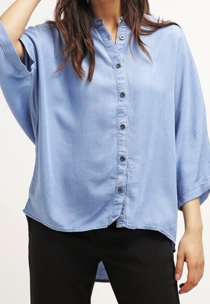 Mujer vestida con una camisa holgada de botones azul claro con las mangas remangadas y pantalones negros, posando frente a un fondo liso y claro.