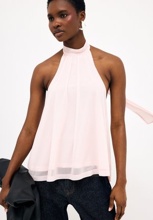 Femme portant un haut dos nu rose pâle, un jean foncé et de grandes boucles d'oreilles blanches, tenant une veste noire pliée dans sa main gauche.