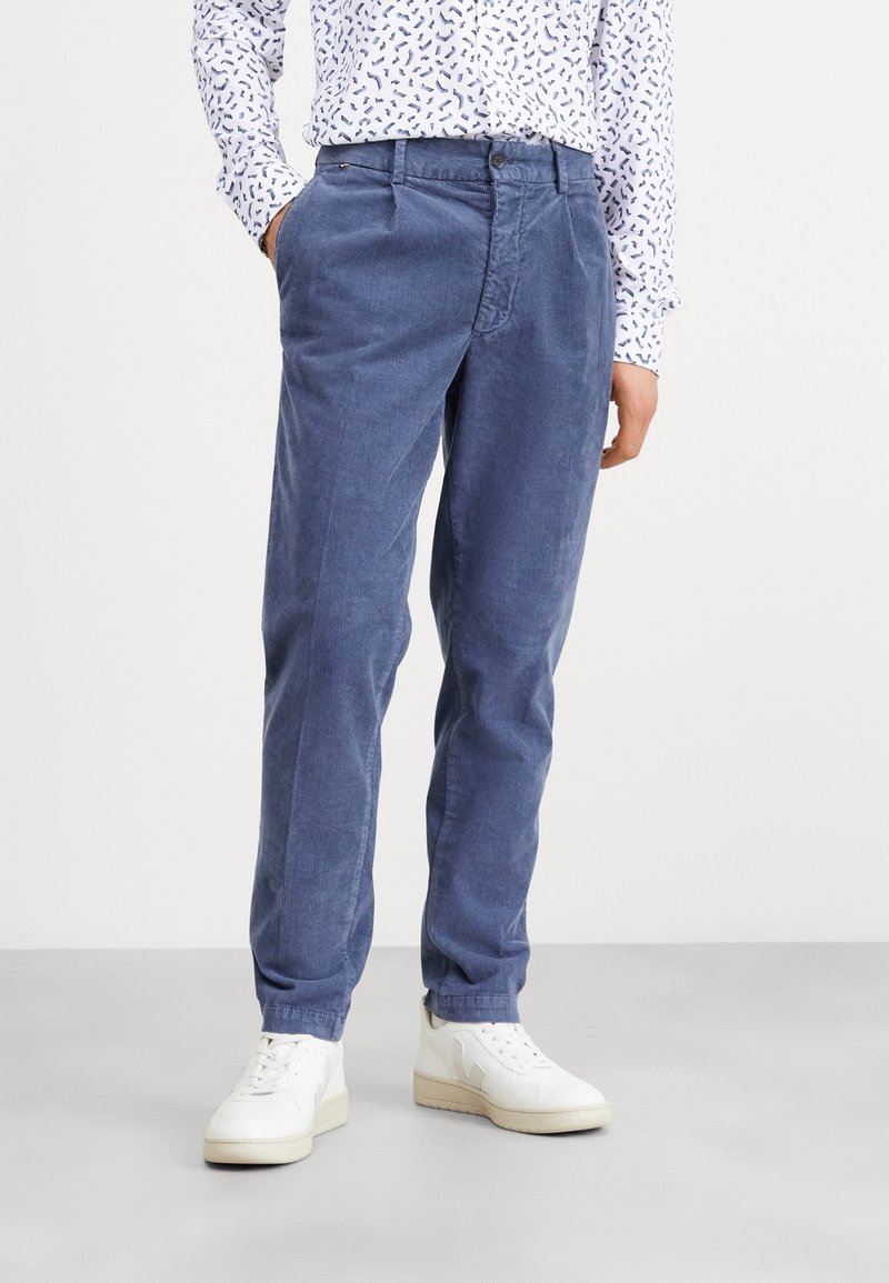 Homme portant un pantalon en velours côtelé bleu, une chemise blanche à motifs et des baskets blanches, debout les mains dans les poches sur un sol gris.
