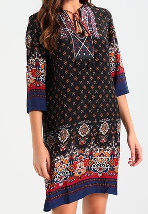 Kobieta w czarnej sukience do kolan z misternymi geometrycznymi i kwiatowymi wzorami w kolorach czerwonym, niebieskim, pomarańczowym i białym oraz z rękawami o długości do łokcia.