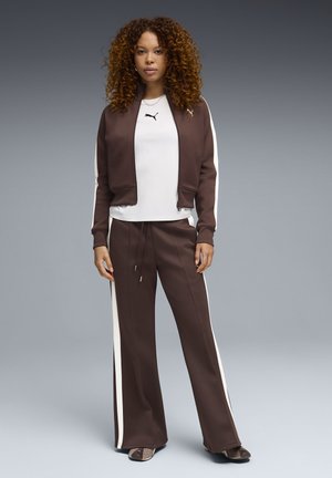 Bruine trainingspak met crème zijstrepen, bestaande uit een bomberjack met ritssluiting en elastische broek met trekkoord. Witte ondershirt en sneakers.