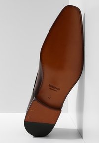 Semelle de chaussure en cuir marron avec une forme arrondie. Présente une couleur fauve, un logo, et de petits éléments métalliques sur le talon. Taille 41 affichée.