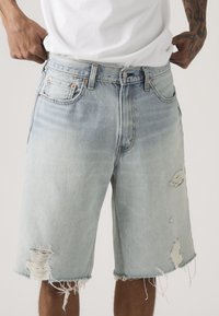 Άνδρας που φοράει ανοιχτό μπλε σκισμένο τζιν σορτς και λευκό μπλουζάκι, ρυθμίζοντας τη μέση με ορατά τατουάζ στα χέρια.