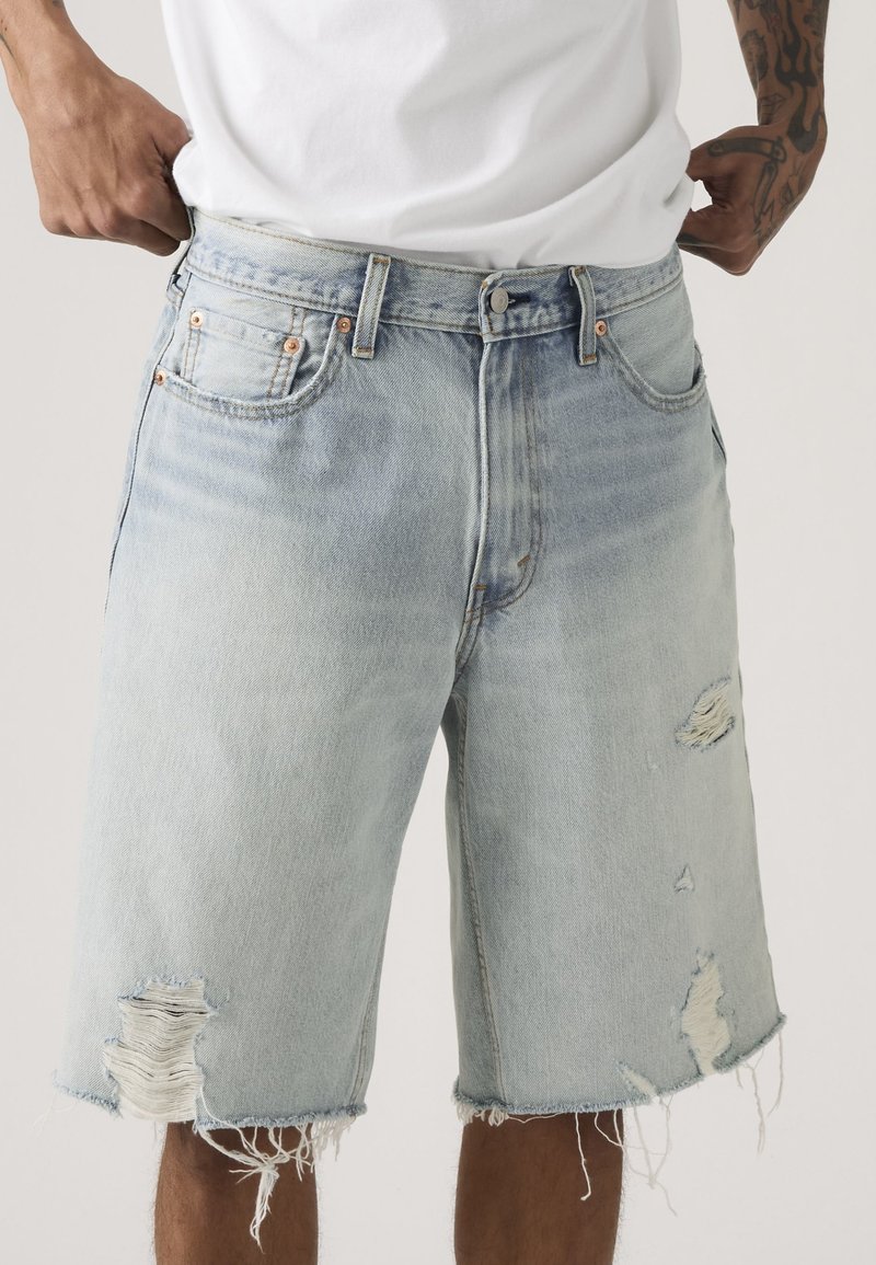 Man in lichtblauwe versleten spijkershorts en een wit T-shirt, die de tailleband verstelt met zichtbare getatoeëerde handen.