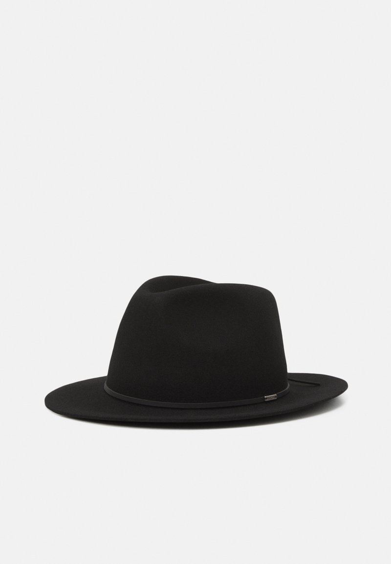 Chapeau fedora noir à large bord avec un fin ruban et une discrète étiquette en métal, présenté sur un fond gris clair uni.