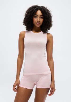 Kvinde med krøllet hår iført en ærmeløs lyserød tætsiddende top og matchende shorts, stående foran en ensfarvet hvid baggrund.