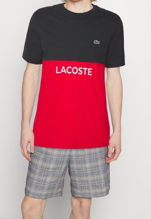 Homme portant un t-shirt Lacoste noir et rouge avec logo et un short à carreaux gris, se tenant devant un fond clair uni.