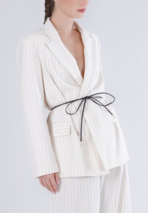 Femme portant un blazer blanc à fines rayures et un pantalon assorti, cintrés à la taille par une fine ceinture noire nouée en nœud.