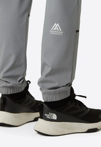 Grå treningsbukser med elastiske mansjetter, med en hvit "Mountain Athletics"-logo. Kombinert med svarte sportssko med "The North Face"-logo.