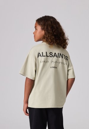 Göndör hajú gyermek oldalra fordulva, bézs ALLSAINTS pólót visel fekete felirattal és japán karakterekkel a hátoldalán.