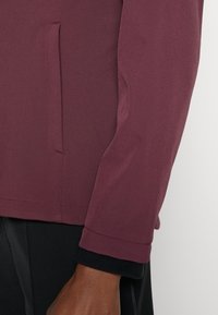 Manche d'une veste bordeaux portée sur un haut à manches longues noir, avec une couture latérale et un ourlet visibles, et une main visible sous le poignet.