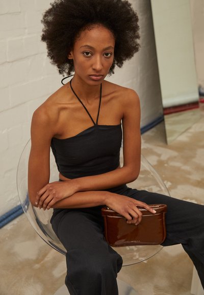 Top preto curto com halter, combinado com calças escuras. Modelo segura uma clutch de couro marrom brilhante enquanto está sentada em uma cadeira de plástico transparente.
