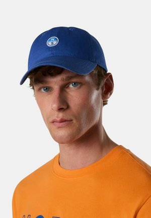 Giovane uomo con occhi azzurri che indossa un berretto blu con un logo a bussola e una maglietta arancione su uno sfondo chiaro uniforme.