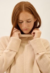 Jasno beżowy sweter dzianinowy z wysokim, prążkowanym kołnierzem, szerokimi prążkowanymi mankietami i teksturowanym środkowym fragmentem. Miękki materiał z subtelnymi detalami.